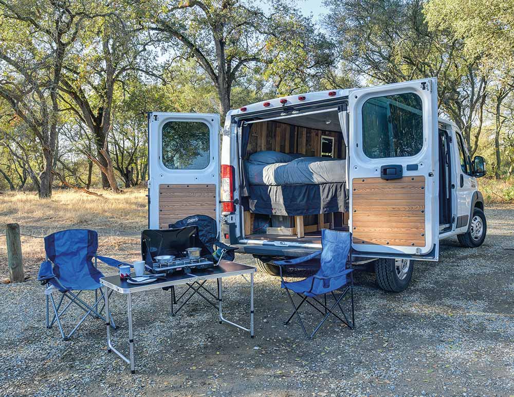 Modified white van open at the back with camping equipment and bed on show