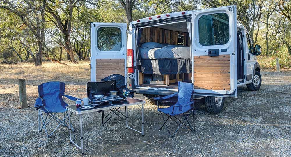 Modified white van open at the back with camping equipment and bed on show