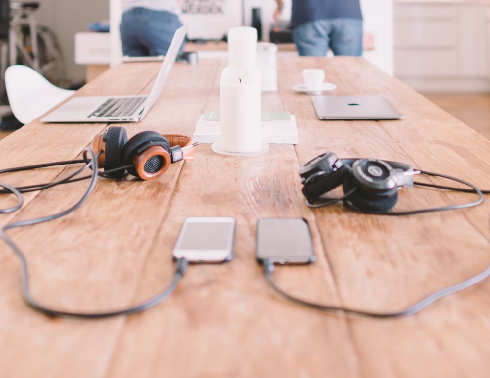 Desk being used for working, with headphones, mobile phones and laptops on the table and people in the background