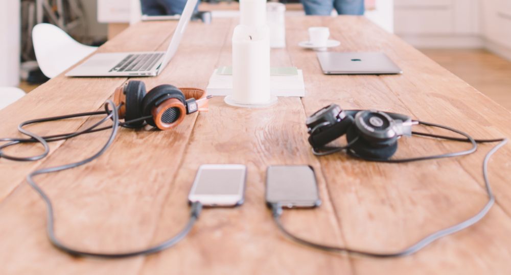 Desk being used for working, with headphones, mobile phones and laptops on the table and people in the background