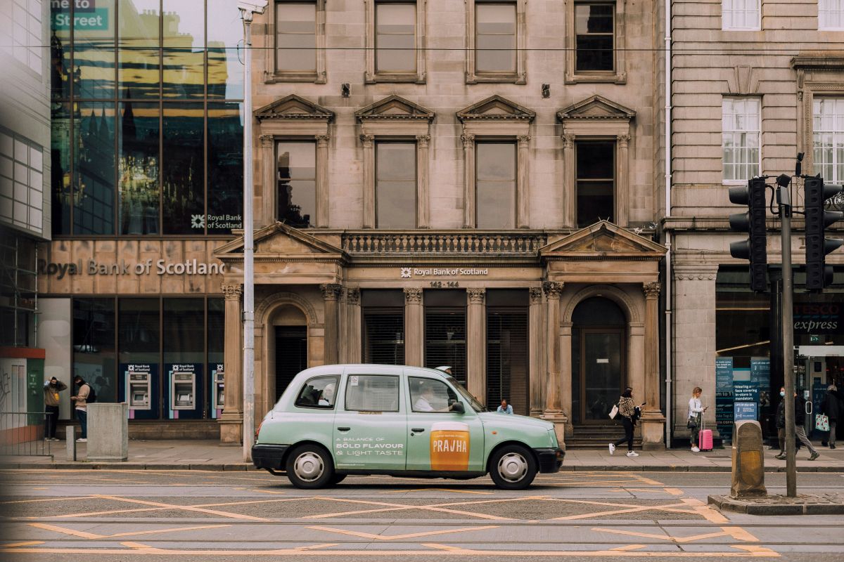 A green taxi drives past the Royal Bank of Scotland building on a city street, where people are walking on the pavement and cash machines are visible