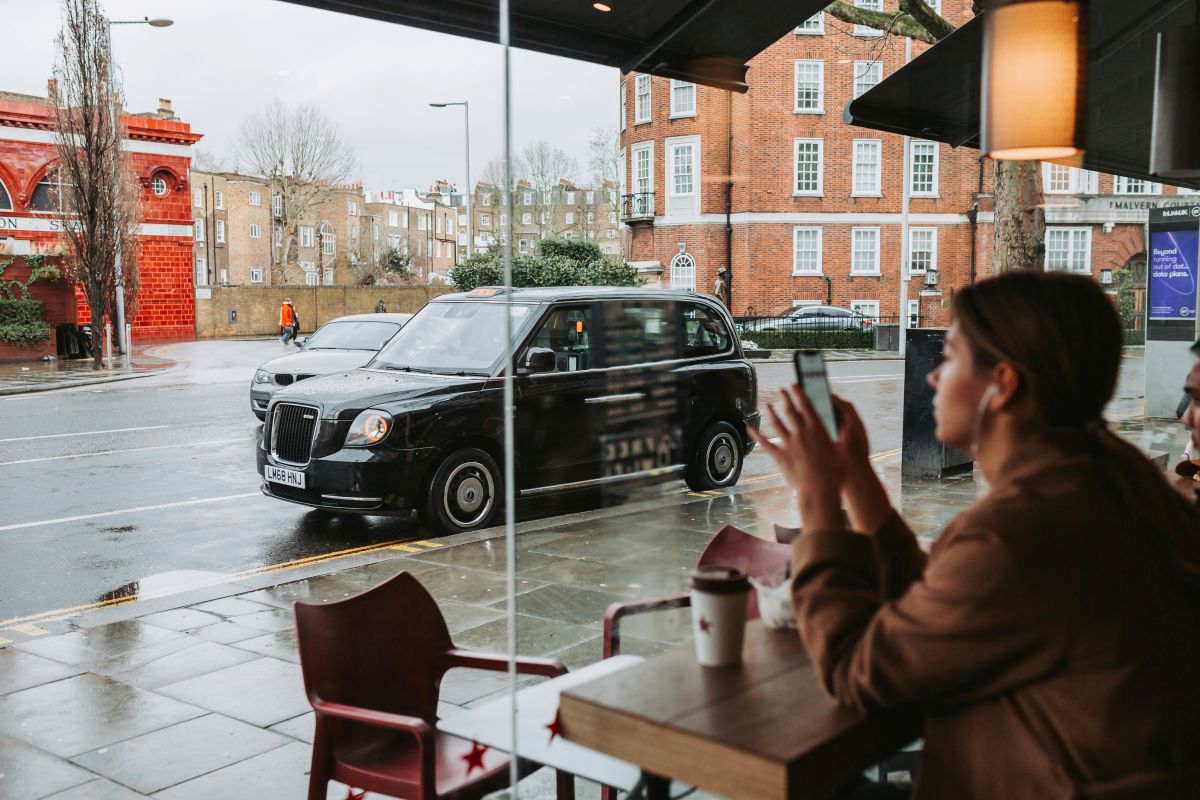 A woman sits at a café window holding her mobile, with a coffee cup on the table. Outside, a black taxi is parked on the rainy street, and red-brick buildings are visible across the road.