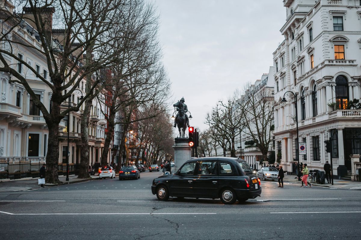 A black taxi drives past a statue of a rider on horseback at a junction lined with trees and white historic buildings on a cloudy day.