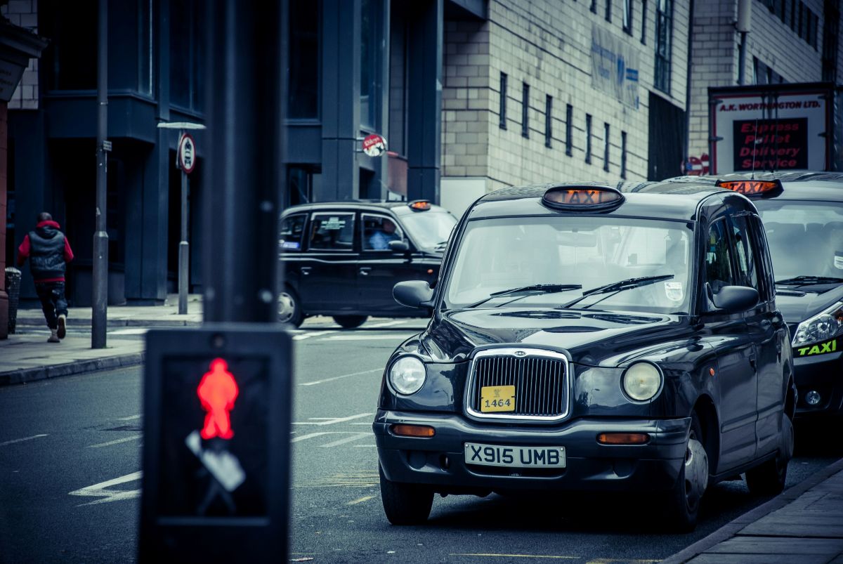 A black taxi waits at a city street junction near a red pedestrian crossing signal, with other taxis and modern buildings in the background.