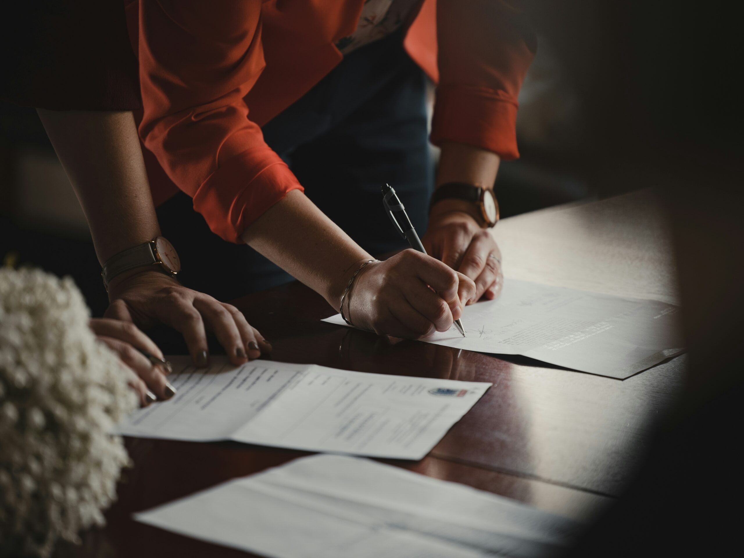A person in an orange jacket leans over a table, signing a document with a pen. An unoccupied chair sits nearby as another person’s hand rests on the table near several sheets of paper, suggesting an official or legal signing.