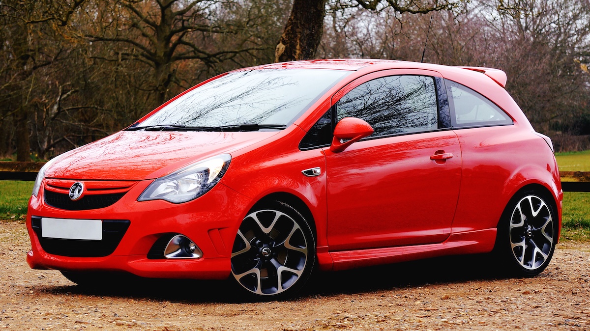 A shiny red compact hatchback car, is parked on a dirt track with trees and grass in the background. The car features black and silver alloy wheels and tinted windows.