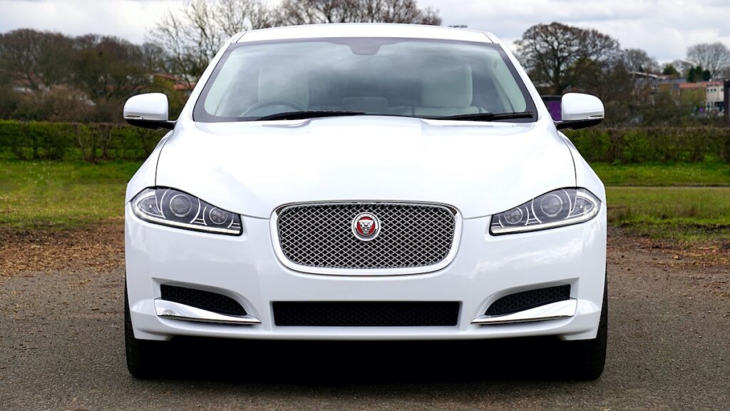 A white Jaguar saloon, is parked on a paved area with grass, trees, and a cloudy sky in the background, seen from the front.