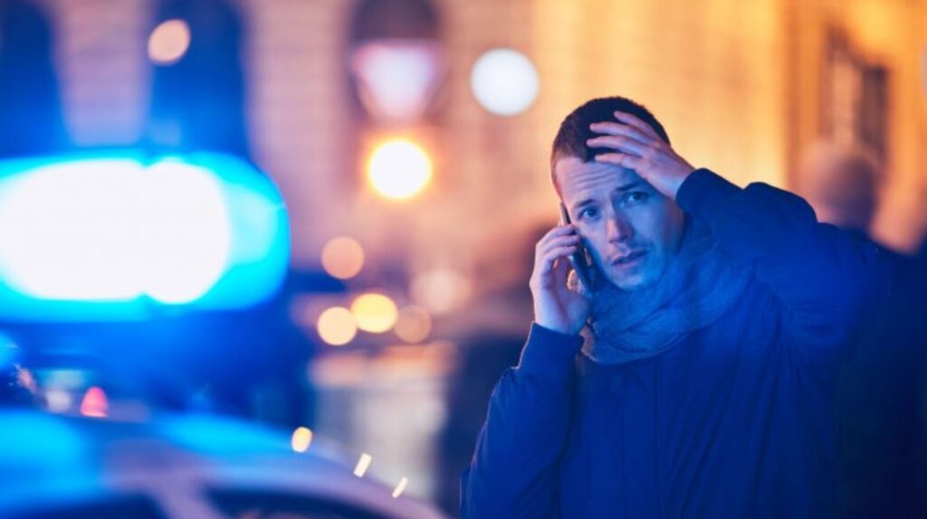Man standing in front of police car on the phone with his hand clutching his head