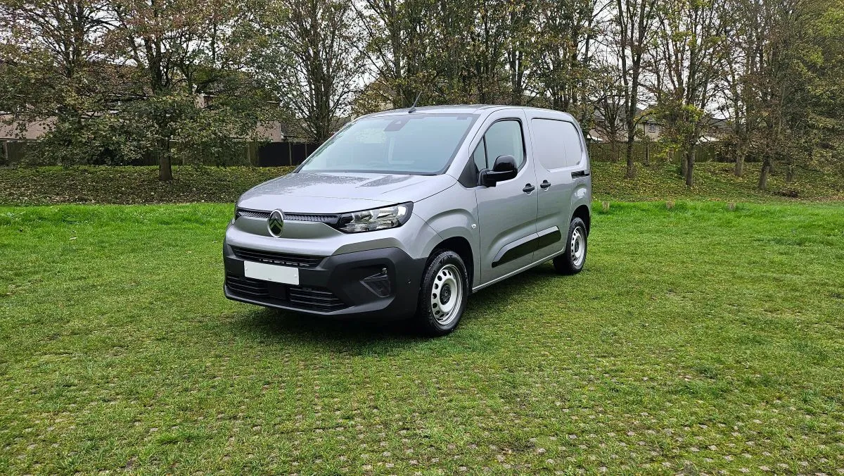 A silver cargo van is parked on a grassy field with trees in the background under a cloudy sky.