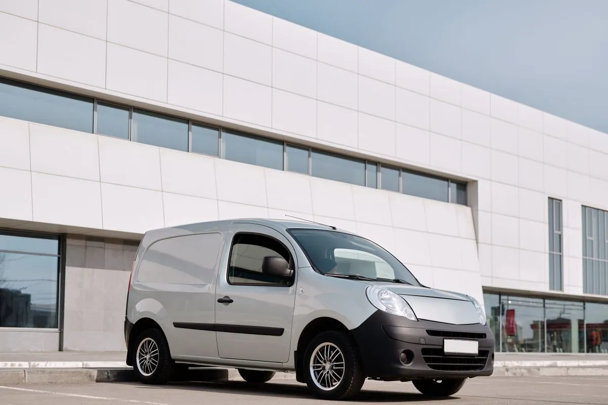 A white delivery van is parked in front of a modern building with large windows and a tiled exterior. The van is unbranded and the sky is clear.