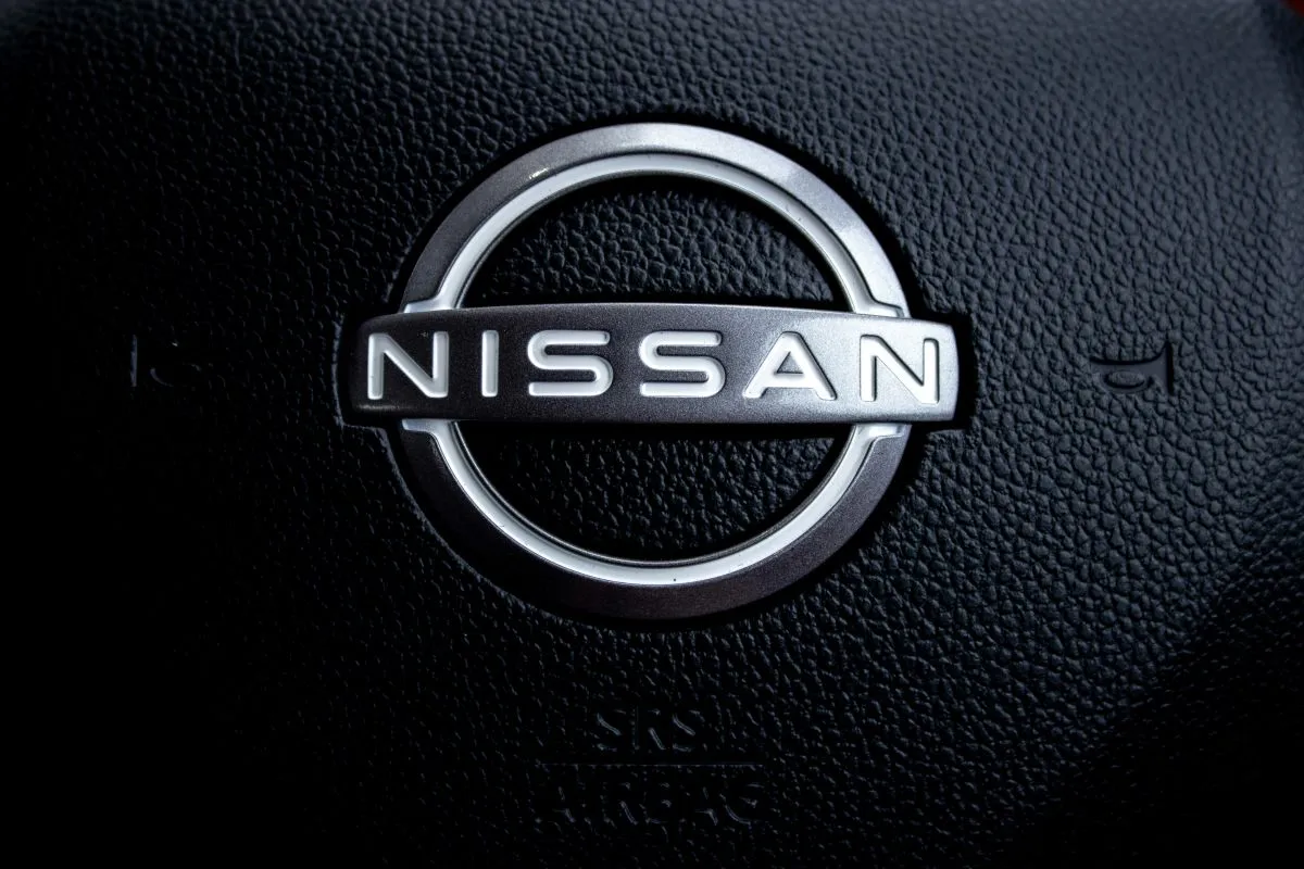 Close-up of a Nissan badge on a black, textured car steering wheel, with part of the SRS airbag label visible below the emblem.