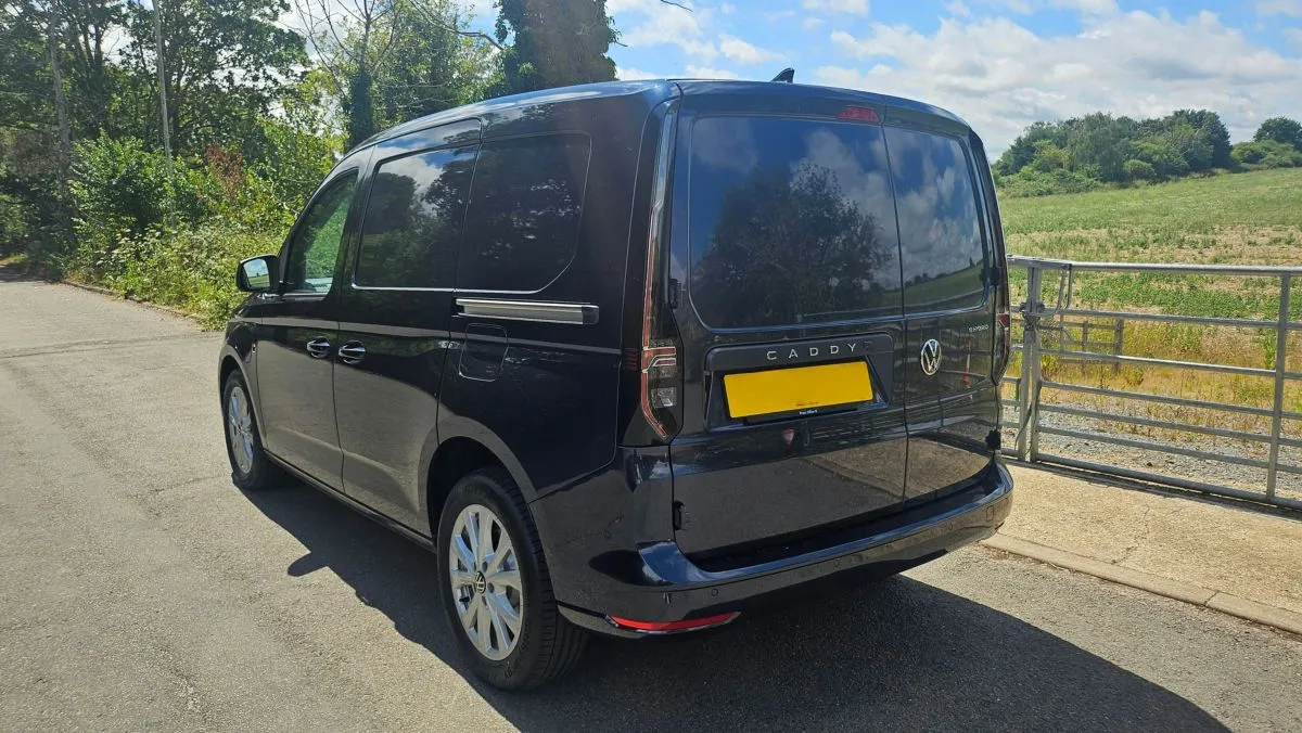 A black Volkswagen Caddy van is parked on a rural road beside a metal gate and grassy fields under a partly cloudy sky.