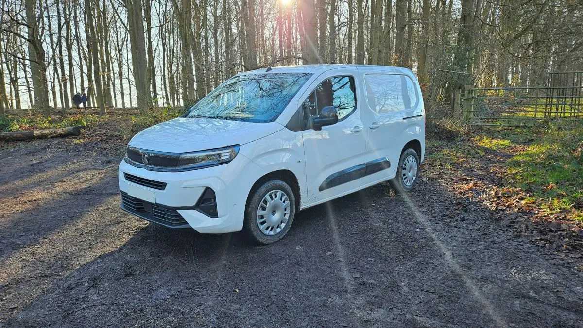 A white van is parked on a sunlit, muddy track in a forested area with bare trees and patches of fallen leaves. Sunlight filters through the trees, casting long shadows.