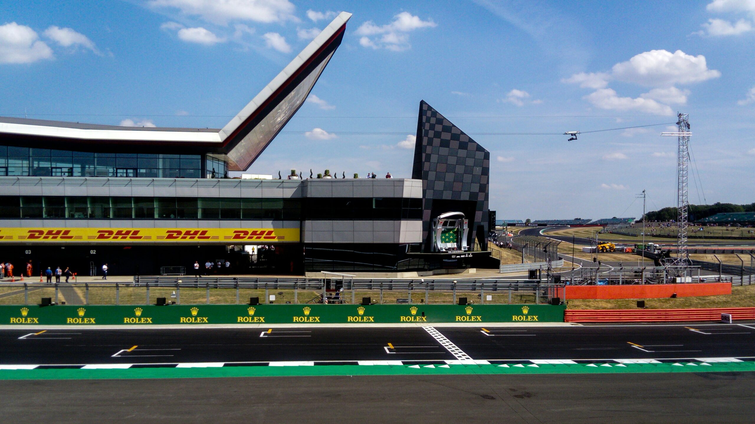 A view of the Silverstone Circuit during the Silverstone Festival, featuring the modern Silverstone Wing building, DHL and Rolex branding, the start/finish line, racetrack, and a blue sky with clouds.