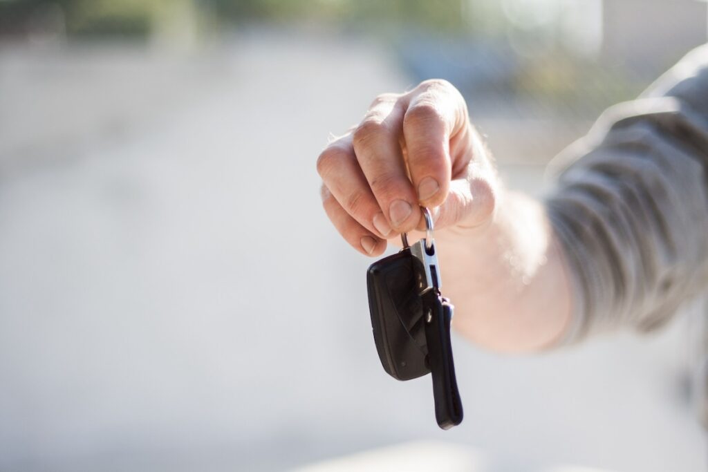 A person’s hand holding out a set of car keys with a black key fob, offering them forward—possibly for a car or quadricycle. The background is blurred and light-coloured.
