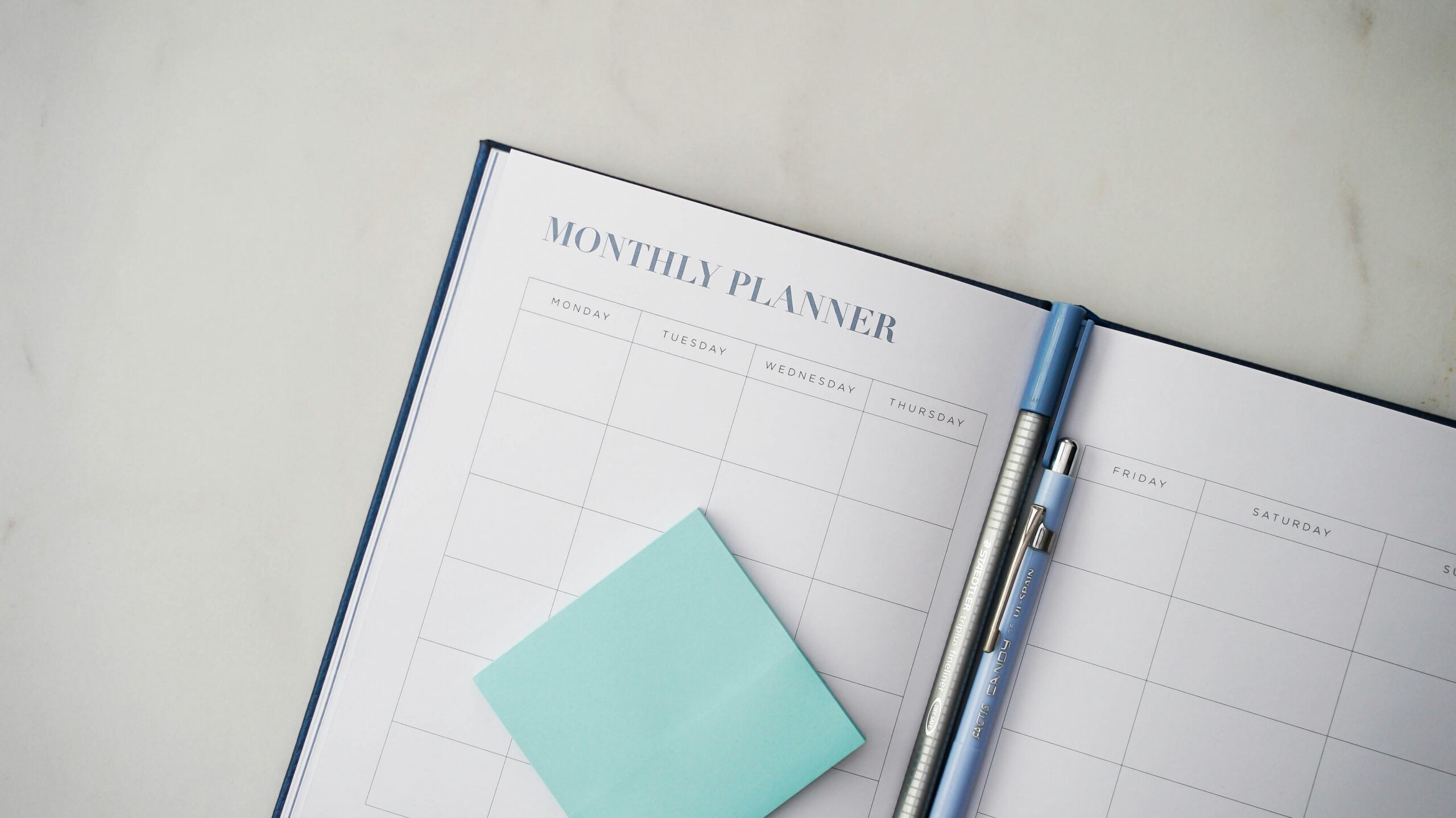 Open monthly diary with blank calendar pages, a blue sticky note, and a blue pen placed on top, all on a white surface—perfect for organising your schedule while living during renovation or planning your next steps in a home remodelling project.