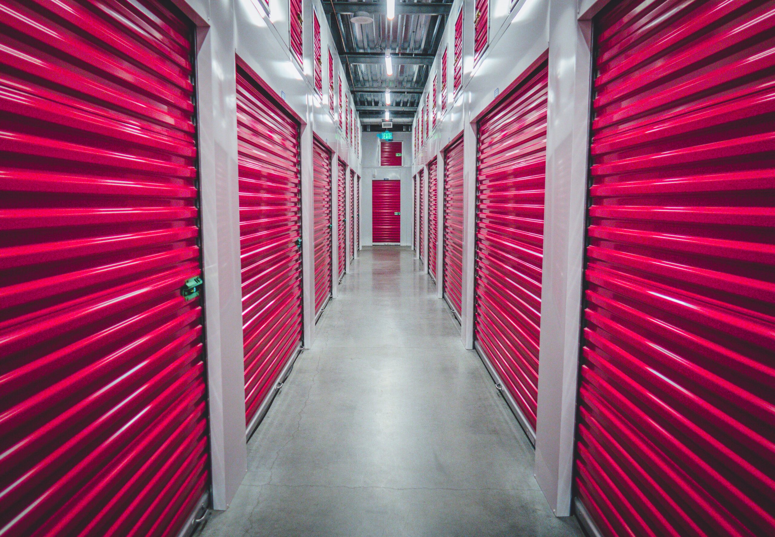 A brightly lit corridor in a storage facility, lined with closed red metal storage unit doors on both sides and a polished concrete floor—an ideal solution if you need extra space while you live in your house during renovation.