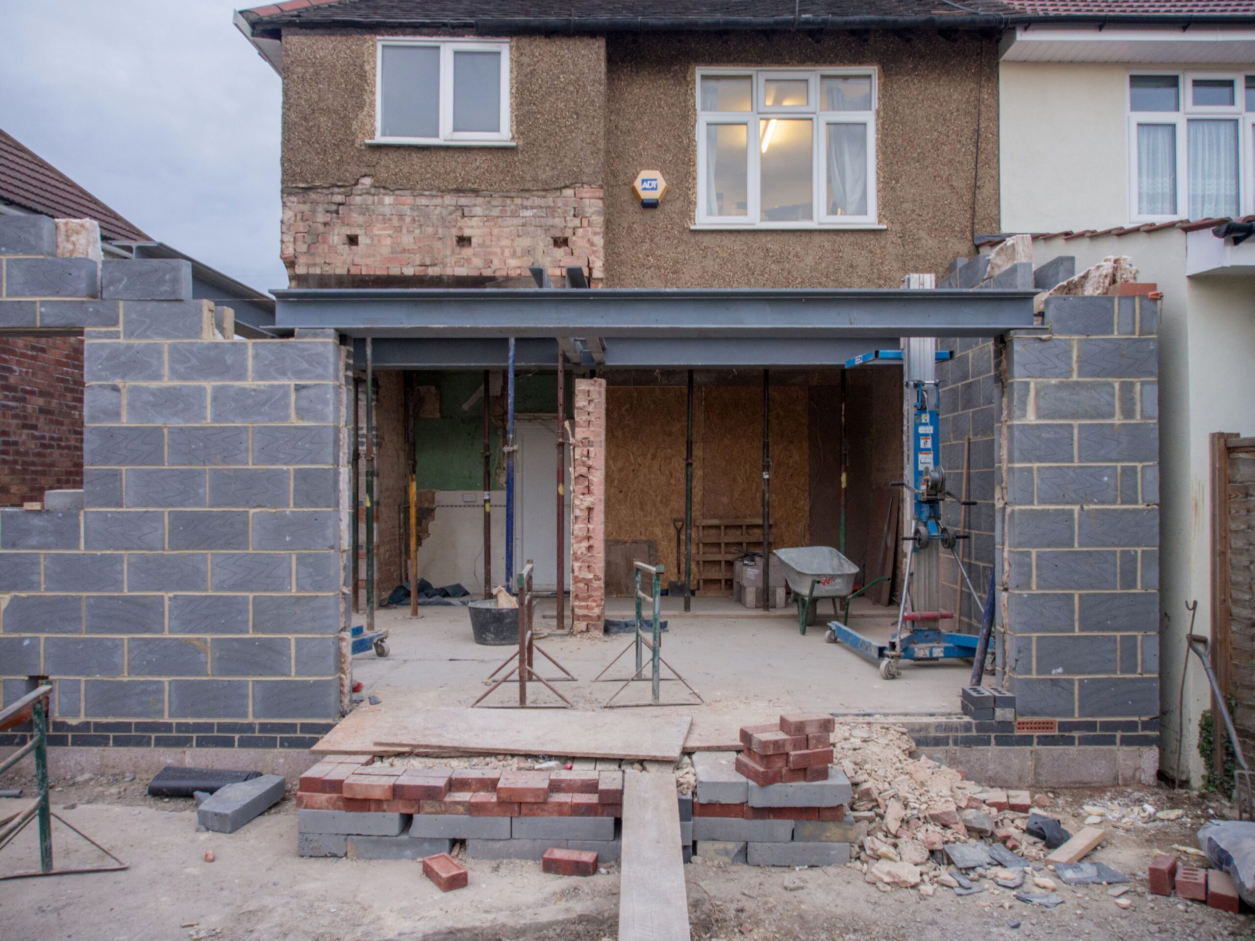 A house undergoing renovation with unfinished brick walls, exposed beams, and building materials such as bricks, scaffolding, and a wheelbarrow on the floor—an authentic snapshot of living in your home during renovation. The upper storey appears complete, offering hope and inspiration for those seeking practical house renovation tips whilst navigating ongoing work.