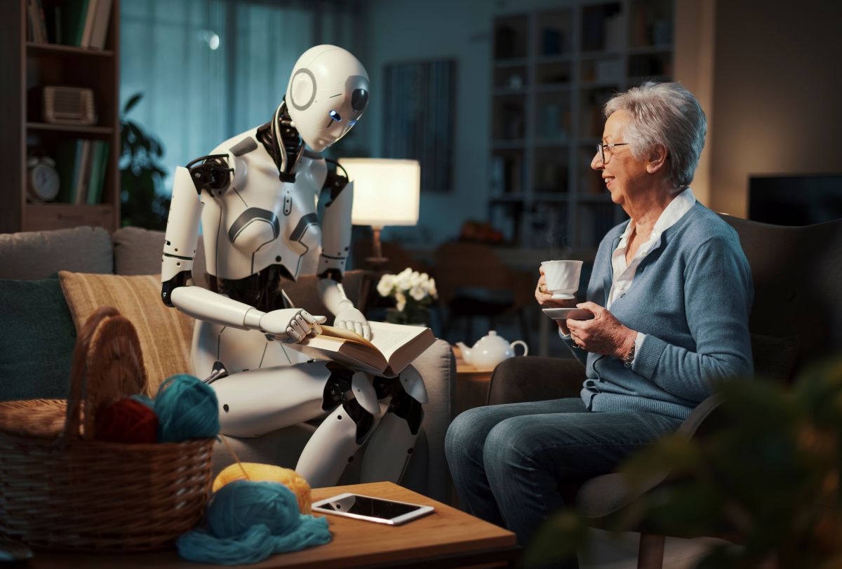 A robot sits on a sofa reading a book with an elderly woman who is holding a cup. They appear to be having a pleasant conversation in a cosy, softly lit living room. A basket of wool and a mobile phone are on the table.